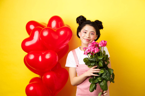 Happy Valentines day. Beautiful asian woman dressed for romantic date, holding bouquet of flowers and smiling. Girl with roses standing near heart balloons, yellow background - 写真・画像