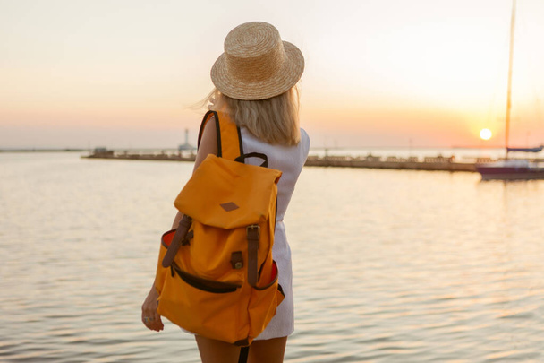 Portrait of a young woman tourist with a large travel backpack at sunrise at sea. Travel and tourism concept - 写真・画像