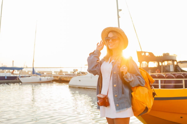 Young woman tourist with large travel backpack and camera at sunrise at yacht Club. Travel and tourism concept - Foto, imagen