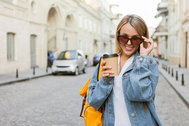 Nice beautiful millennial tourist with backpack in jeans jacket holds coffee cup and discover European city at holiday travel. - Фото, изображение