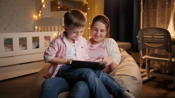 幼い息子と一緒に笑顔の母親は、夜に豆袋に座って、タブレットコンピュータを歌う。夜間に一緒に時間を持つ子供の教育と家族の概念 - 映像、動画