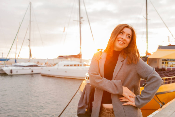 Egy fiatal, vidám, őszi kabátos nő portréja, aki egy jachtklubban pózol napkeltekor. - Fotó, kép