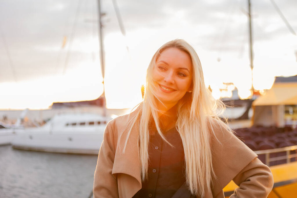 Portrait of young cheerful woman in autumn coat posing in a yacht club at sunrise - Photo, image