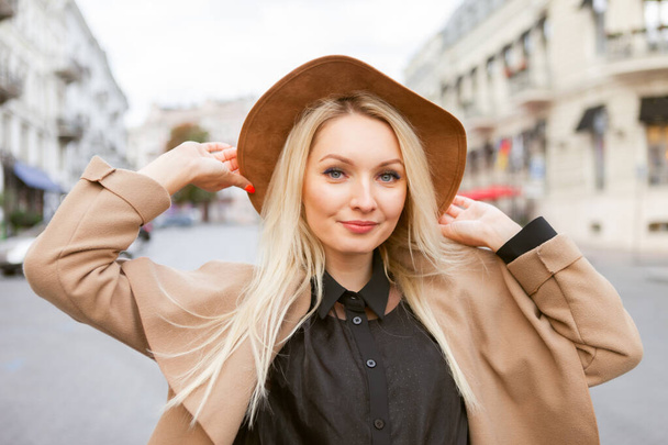Portret młodej, stylowej blondynki w jesiennym płaszczu i kapeluszu w europejskim mieście. Piękna atrakcyjna dziewczyna - Zdjęcie, obraz