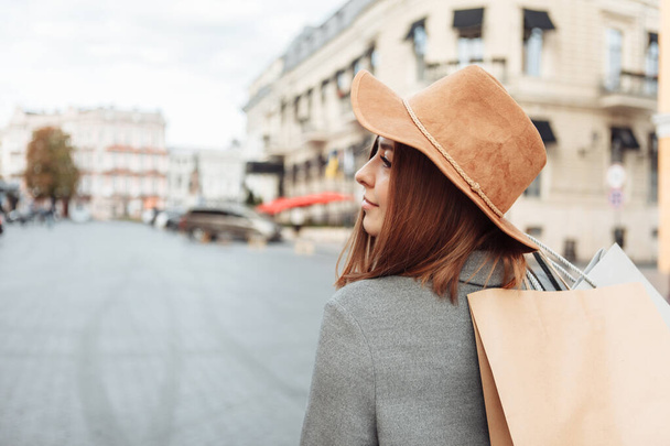 Cute attractive shopaholic funny woman in autumn coat and hat holding many paper shopping bags iin European city - Фото, изображение