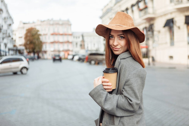 Stylish millennial woman in a slicker coat and felt hat jerks a coffee cup on the go while walking in a european city - Photo, Image