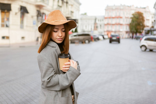 Stylish millennial woman in a slicker coat and felt hat jerks a coffee cup on the go while walking in a european city - Photo, Image