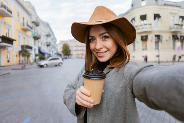 Stylový tisíciletá žena v pláštěnce a plstěný klobouk škubne šálek kávy na cestách a dělá selfie v evropském městě - Fotografie, Obrázek