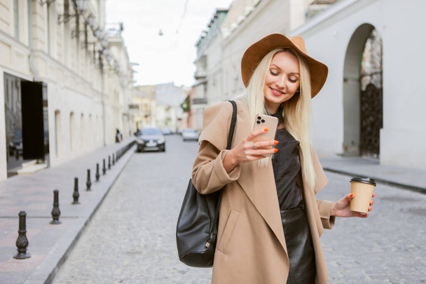 Joven mujer rubia milenaria vestida con abrigo y sombrero de otoño utiliza teléfono inteligente y sostiene una taza de café sobre la marcha en una ciudad europea. - Foto, Imagen