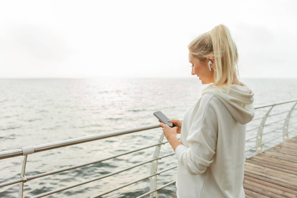 Молодая блондинка в белой толстовке пользуется смартфоном и слушает музыку на наушниках на пляже - Фото, изображение