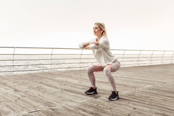 Jeune femme en forme entraîne les jambes avec des élastiques de fitness sur la plage. Mode de vie sain - Photo, image