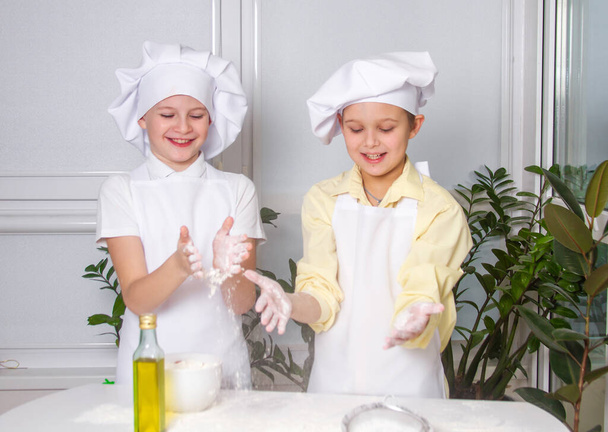 Dos lindos chicos europeos vestidos como cocineros están amasando harina, leche, los niños están jugando en la cocina, los adolescentes están haciendo un pastel con la masa. - Foto, Imagen