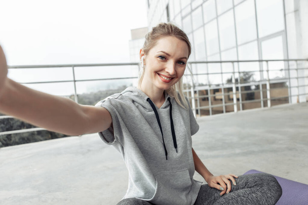 Αυτοπροσωπογραφία. Fitness γυναίκα ακούγοντας μουσική σε earphiones και χαλαρώνοντας, ενώ κάθεται στο χαλάκι στην οροφή του κτιρίου. Αστικό στυλ - Φωτογραφία, εικόνα