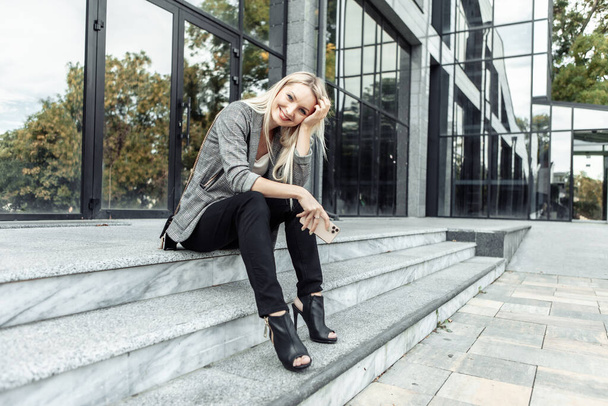 Glückliche Geschäftsfrau sitzt in der Pause auf den Stufen und hält ihr Smartphone vor den Hintergrund des Business Centers - Foto, Bild