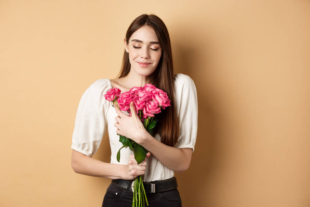 Walentynki. Czuła i romantyczna dziewczyna, pachnąca różami i uśmiechnięta z zamkniętymi oczami. Dziewczyna przytulając prezent kwiaty od kochanka, stojąc na beżowym tle - Zdjęcie, obraz