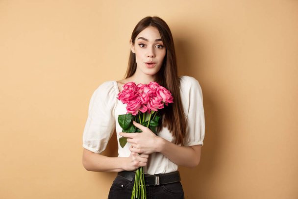 Valentines day. Image of surprised girlfriend thanking for flowers, receive pink roses from lover and looking grateful at lover, standing on beige background - Fotografie, Obrázek