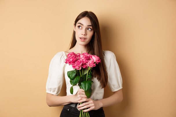 Romantický Valentýnský koncept. Zasněná žena s kyticí růžové růže, dívá se stranou na logo, drží květiny na béžovém pozadí - Fotografie, Obrázek