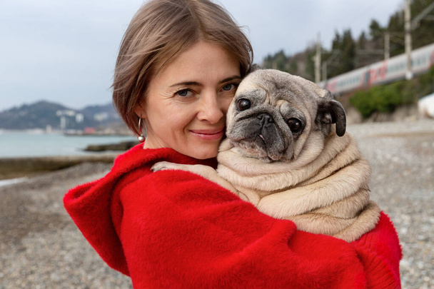 A pug in the arms of a woman in a red coat. Warm winter day by the sea. - Foto, imagen