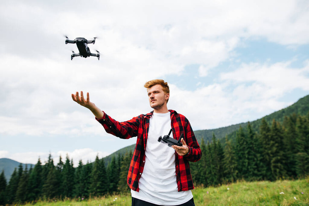 L'uomo in camicia rossa vola su un drone durante un'escursione in montagna, si erge su un prato con un telecomando in mano e guarda il gadget. Blogger crea video di viaggio dalle montagne. - Foto, immagini