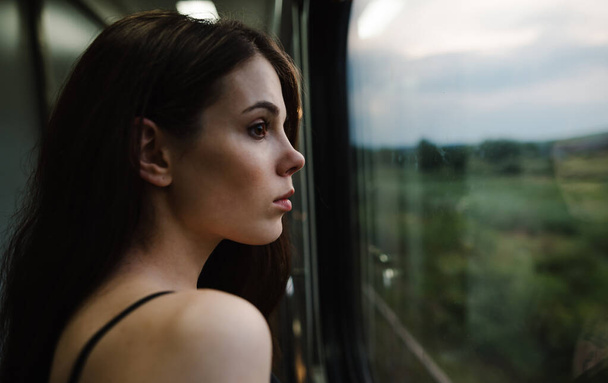 Close-up portrait of an attractive female train passenger standing by the window and looking at the beautiful road scenery. Dreamy lady looks out the train window and thinks. - Fotó, kép