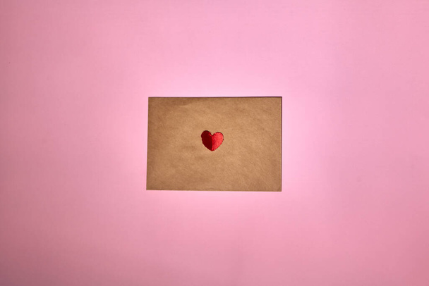 A red heart lies on a brown envelope for Valentine's Day. Pink backdrop with envelope and red heart. Love letter or message concept. Flat-lay, top view. Copy space for your text.  - 写真・画像
