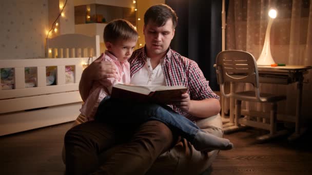 Genç baba küçük oğluyla yatak odasında oturup masal kitabı okuyor. Çocuk eğitimi ve aile kavramının geceleri birlikte vakit geçirmesi - Video, Çekim