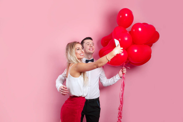 Valentines day. Romantic couple taking selfie on mobile phone and standing with red balloons on pink background - Photo, image