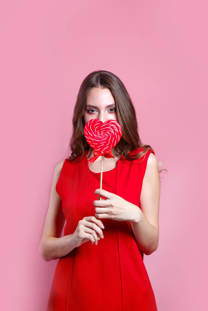 Beautiful brunette woman in red dress holding a red heart-shaped lollipop on pink background. Valentines Day - Fotografie, Obrázek