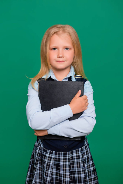 Little schoolgirl with a book in her hands and a backpack on her back posing on a green background. - Zdjęcie, obraz