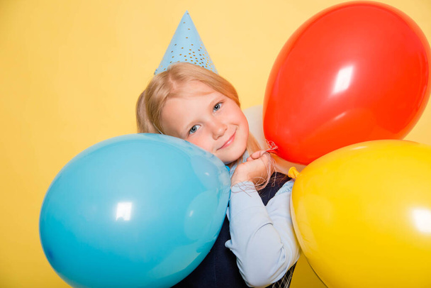A girl in a school uniform with a festive cap on her head is holding balloons in her hands isolated on a yellow background. - Photo, image