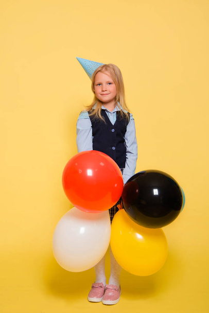 A girl in a school uniform with a festive cap on her head is holding balloons in her hands isolated on a yellow background. - Fotoğraf, Görsel