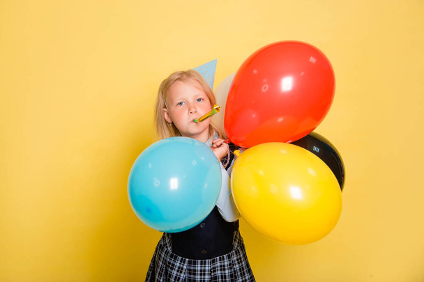 Ένα κορίτσι με σχολική στολή και ένα εορταστικό κώνο στο κεφάλι της κρατά μπαλόνια στα χέρια της και φυσάει ένα σωλήνα που απομονώνεται σε κίτρινο φόντο. - Φωτογραφία, εικόνα
