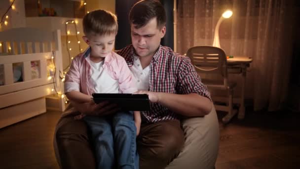 若い笑顔の父親がインターネットを閲覧し、夜に寝室で彼の小さな息子と漫画を見ての肖像画。夜間に一緒に時間を持つ子供の教育と家族の概念. - 映像、動画