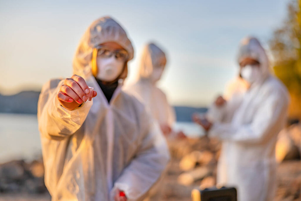 Ученый в белом защитном костюме держит образец воды на пляже - Фото, изображение