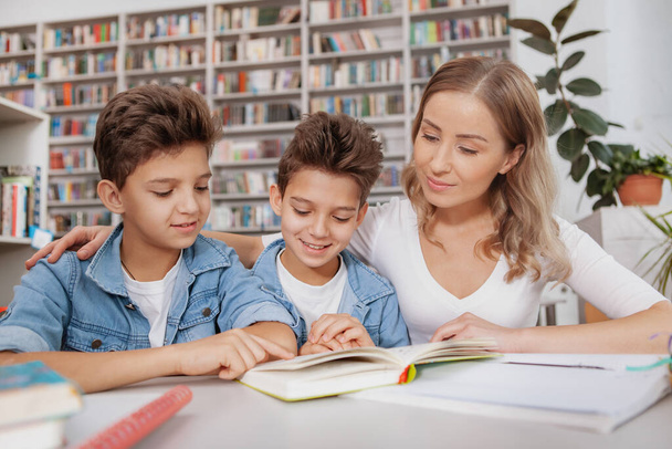 Beautiful woman helping her twin sons with homework, reading a book at the library. Two little brothers studying with their mom - Фото, изображение