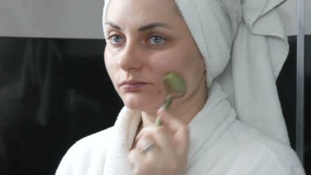 Kaunis nainen, jolla on pyyhe päässä tekemässä hierontaa jade-rullalla kiven kasvoille kylpyhuoneessa. Kiinalaisia kauneustyökaluja. Imusolmukkeiden salaojitus ihon hieronta vastaan ikääntyminen ryppyjä - Materiaali, video