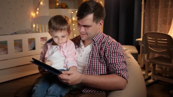 クリスマスのために飾られた寝室でタブレットコンピュータを使用して父親と笑顔の少年のパンニングショット。夜間に一緒に時間を持つ子供の教育と家族の概念. - 映像、動画