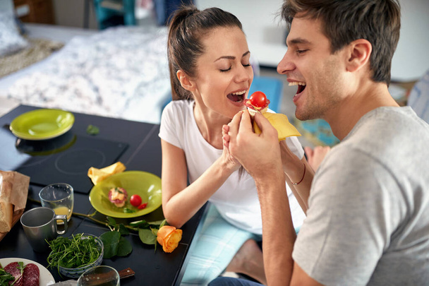 A young couple having fun while feeding each other during a breakfast in a cheerful atmosphere at home. Couple, home, breakfast, togetherness - Photo, Image