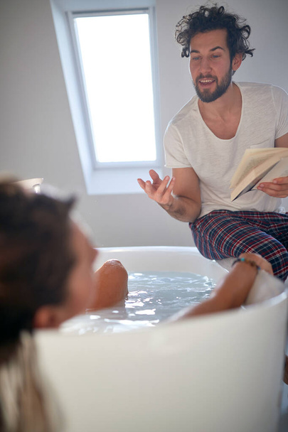 A young man who got into the role while reading a book to his girlfriend who enjoying the bath in the relaxed atmosphere of the bathroom. Love, relationship, together - Foto, afbeelding
