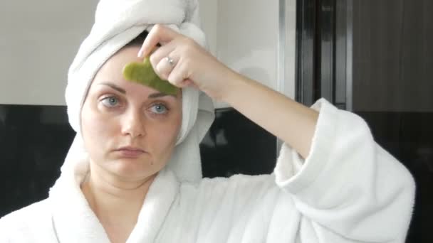 Bella donna con asciugamano sulla testa facendo massaggio con un raschietto di pietra di giada per guazzo fronte Massaggio in bagno. Strumenti di bellezza cinesi. Massaggio linfatico drenante cutaneo contro l'invecchiamento delle rughe - Filmati, video