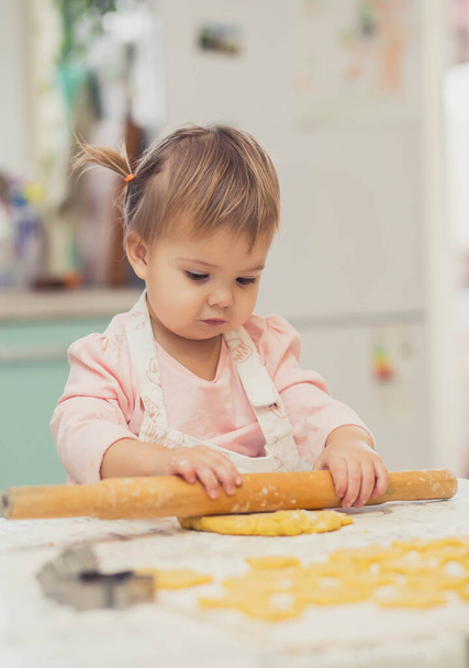 Adorable baby in an apron is rolling out the dough in the kitchen - Photo, Image