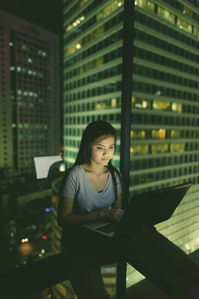 Portrait de jeune belle femme asiatique utilisant un ordinateur portable contre fenêtre en verre avec vue sur la ville la nuit - Photo, image