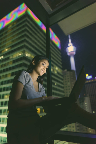 Портрет молодої вродливої азіатки, що використовує ноутбук проти скла, з видом на місто вночі. - Фото, зображення