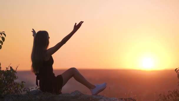 Fiatal elegáns nő fekete rövid ruhában ül egy sziklán pihentető szabadban nyáron este. Divatos nő élvezi a meleg naplementét a természetben. - Felvétel, videó
