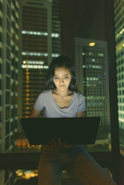 Портрет молодой красивой азиатки с ноутбуком у стеклянного окна с видом на город ночью - Фото, изображение