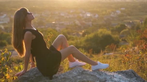 Jonge elegante vrouw in zwarte korte jurk zittend op een rots ontspannen buiten in de zomer avond. Modieuze vrouw geniet van warme zonsondergang in de natuur. - Video
