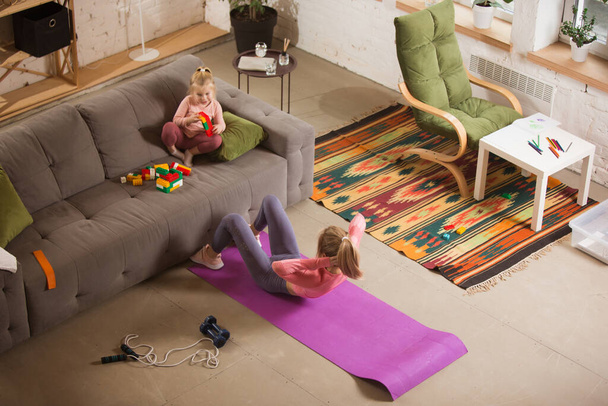 Young woman exercising fitness, aerobic, yoga at home, sporty lifestyle. Getting active while her child playing on the background, home gym. - Foto, Imagen