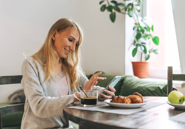 Beautiful blonde young woman having breakfast and using mobile in living room at the home - 写真・画像