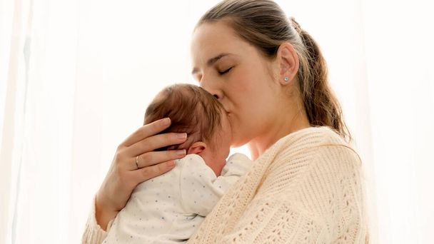 Closeup portrait of young loving mother kissing her little newborn baby at window - Photo, Image