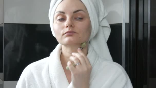 Bella donna con asciugamano sulla testa che fa un massaggio con un rullo di giada per il mento del viso di pietra in bagno. Strumenti di bellezza cinesi. Massaggio linfatico drenante cutaneo contro l'invecchiamento delle rughe - Filmati, video
